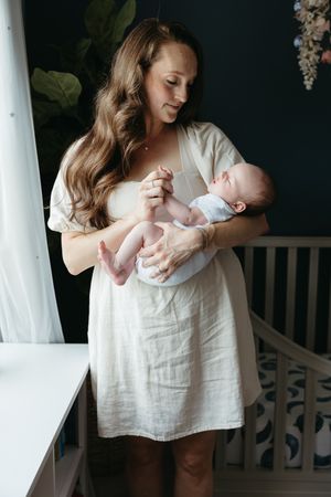 A tender moment in a nursery as a woman in a white dress holds and gazes lovingly at a newborn baby.