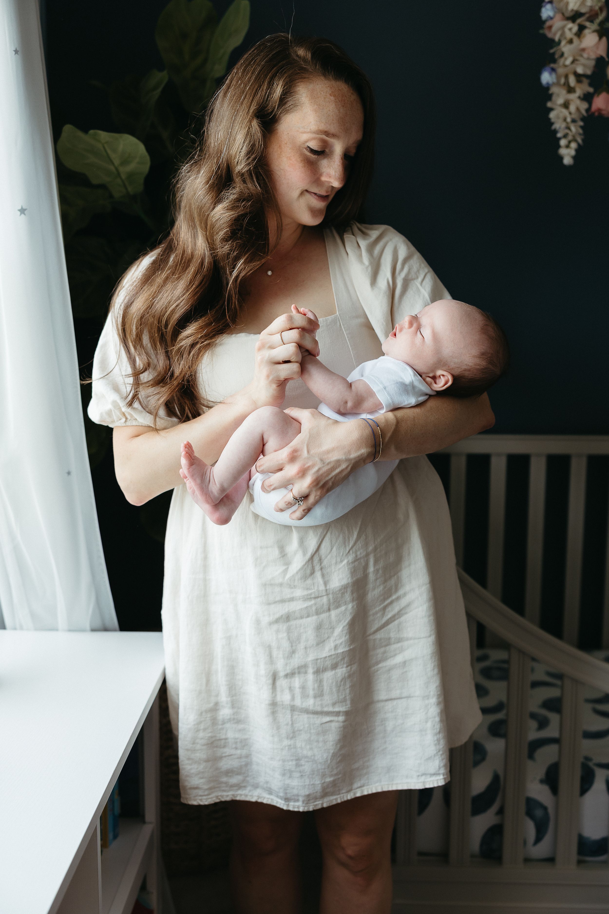 A tender moment in a nursery as a woman in a white dress holds and gazes lovingly at a newborn baby.