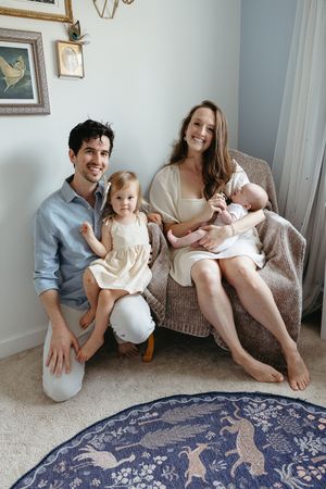 A family of four sits together in a cozy corner with a decorative blue rug and framed artwork on the wall.