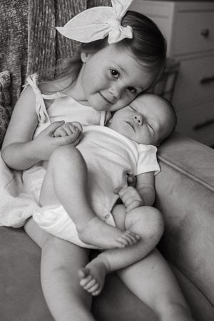 A black and white portrait of siblings cuddling together, with the older child wearing a white bow and a sweet smile.