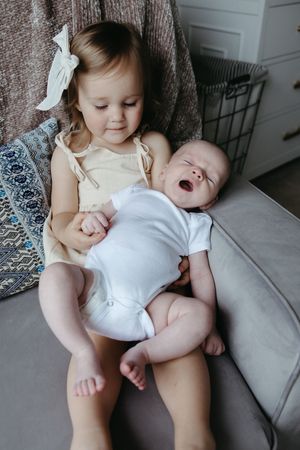 A young child in a cream dress sits on a couch holding a laughing baby in a white onesie.