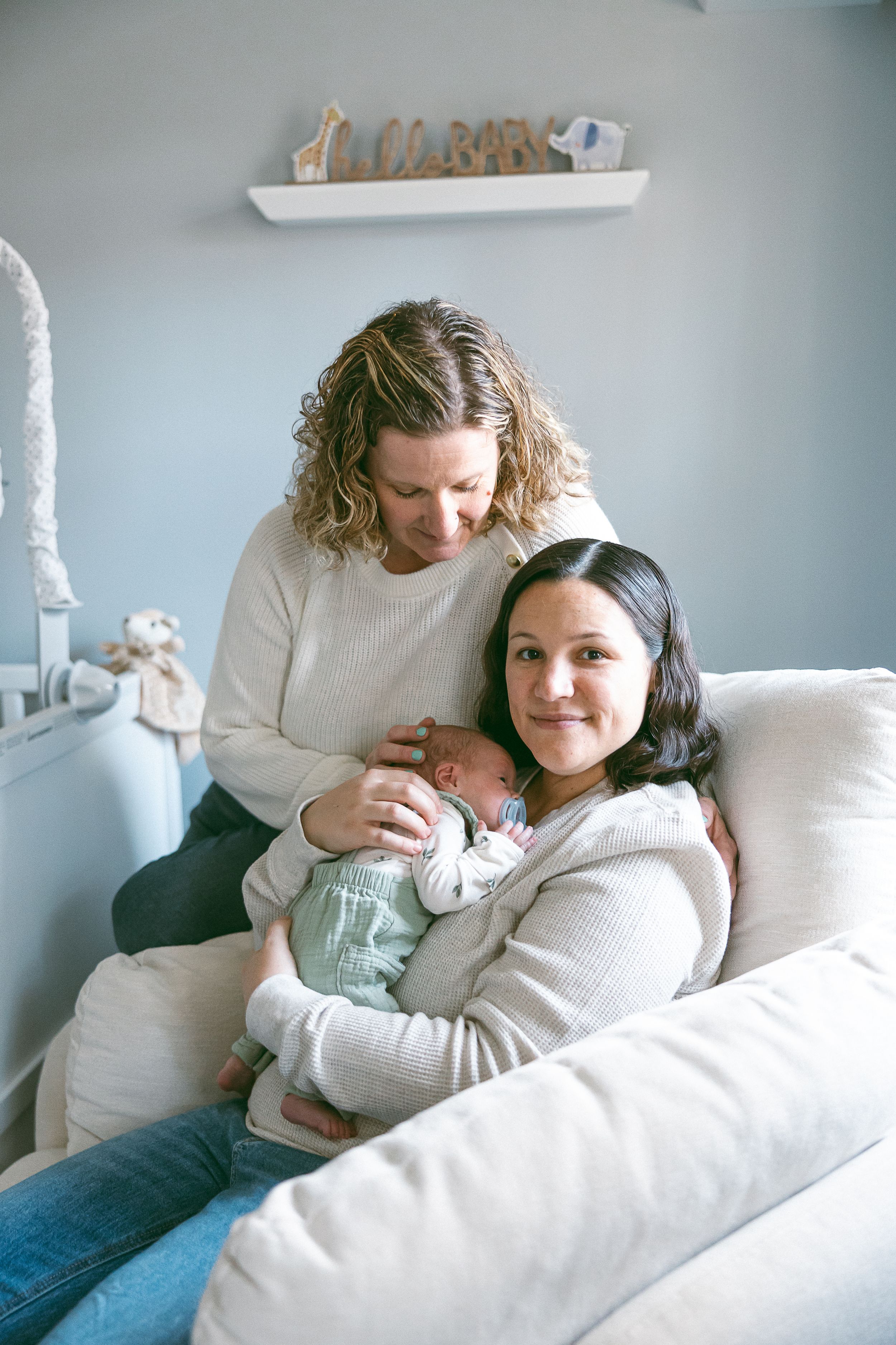 Two adults sit on a white couch with a newborn baby in a cozy living room with light blue walls.