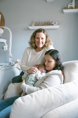 Two people sitting on a white couch with an infant in a bright, cozy room with wall shelves.