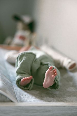 Close-up view of tiny baby feet in sage green pants resting on soft gray fabric.