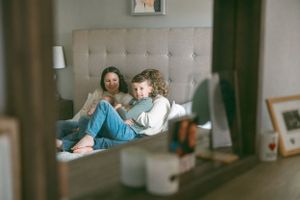 A parent and child relax together on a grey upholstered bed while looking at a mobile device.