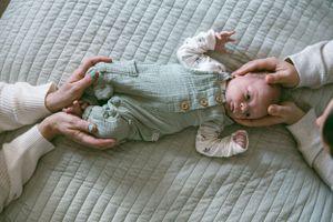 Baby in gray striped overalls lies on quilted bedspread while adult hands reach in to offer comfort.