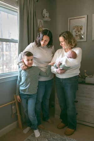 A family of four stands together in a cozy nursery room by a window with soft natural lighting streaming in.