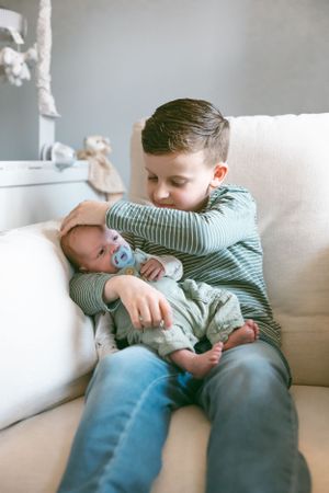 Sibling love displayed on white couch as older child in striped shirt gently holds sleeping baby with pacifier.