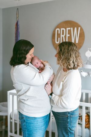 Two people in white sweaters admire a newborn baby in a neutral-toned nursery with a CREW sign on the wall.