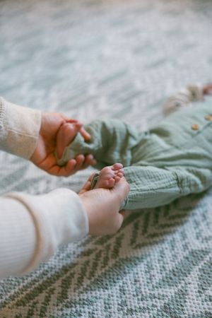Close-up view of hands holding tiny feet of an infant wearing soft sage green clothing on a textured blanket.