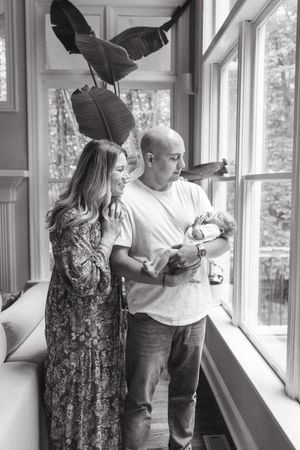 Black and white photo of a couple with their newborn baby standing by sunlit windows with large tropical plants.