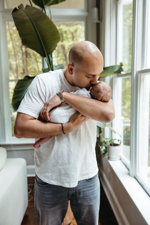 A tender moment between a parent and newborn baby by a sunny window with a large houseplant in the background.