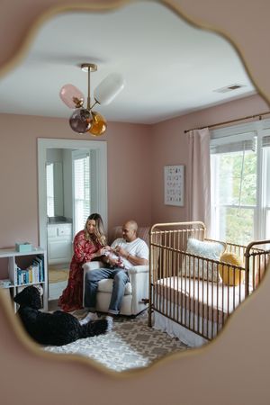 Modern nursery room with pink walls, gold crib, patterned rug and hanging pendant light fixture viewed through decorative mirror.
