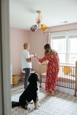 Two people standing in a nursery room with pink walls and a dog next to them while looking at something together.