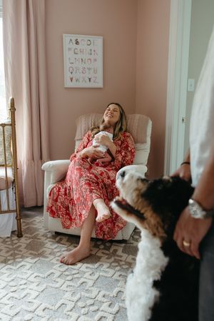 A person in a red floral dress relaxes in a nursery armchair while two dogs watch from the doorway.
