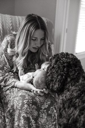 A tender moment between a newborn baby and a large black dog while a woman in a floral dress looks on in black and white.