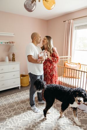 A couple shares an intimate moment in a nursery room with two dogs while standing on a patterned area rug.
