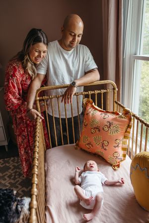 Parents standing over a wooden crib with their newborn baby while decorative orange pillows adorn the nursery.