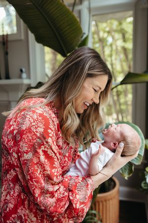 A woman in a red floral dress shares a tender moment with a baby near a houseplant in natural lighting.