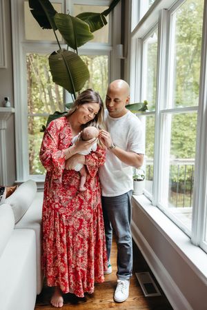 Parents in a bright room with large windows care for their newborn baby near a decorative indoor plant.