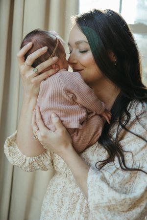A tender moment between mother wearing lace dress and baby in pink knit sweater by window in soft natural light.