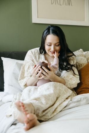 Relaxed person wearing white lace loungewear smiles while using phone in cozy bed with white linens and framed wall art.