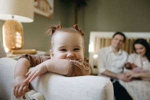 A young child smiles while leaning on a white couch as family members sit in the background.