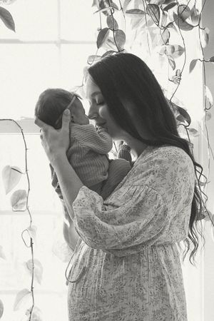 A tender black and white photograph showing a baby being lovingly held near a window with trailing vines in soft light.