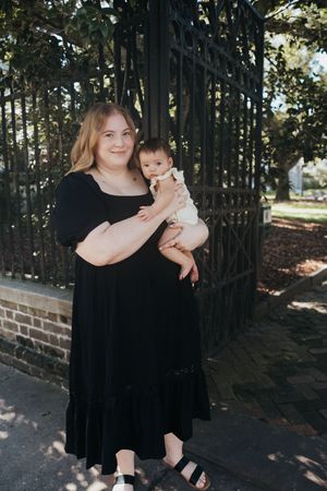 A parent in a black dress holds a baby in white clothing while standing near a brick wall and wrought iron fence.