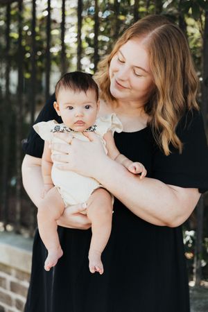 A loving parent gazes down while holding a baby wearing a white outfit against an outdoor garden backdrop.