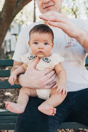 Baby wearing embroidered white outfit sitting on caregiver's lap outdoors on park bench.