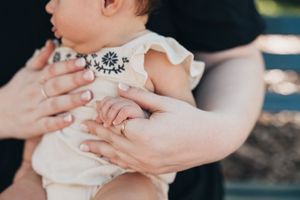 Parent's hands tenderly hold a baby wearing a white embroidered outfit, showing a close-up of their embrace.