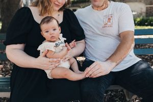 Parent holds baby wearing white onesie while sitting together outdoors during casual family photography session.