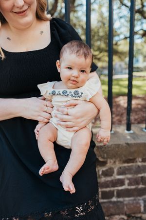Adorable baby wearing a white onesie sitting in someone's lap during an outdoor photoshoot.