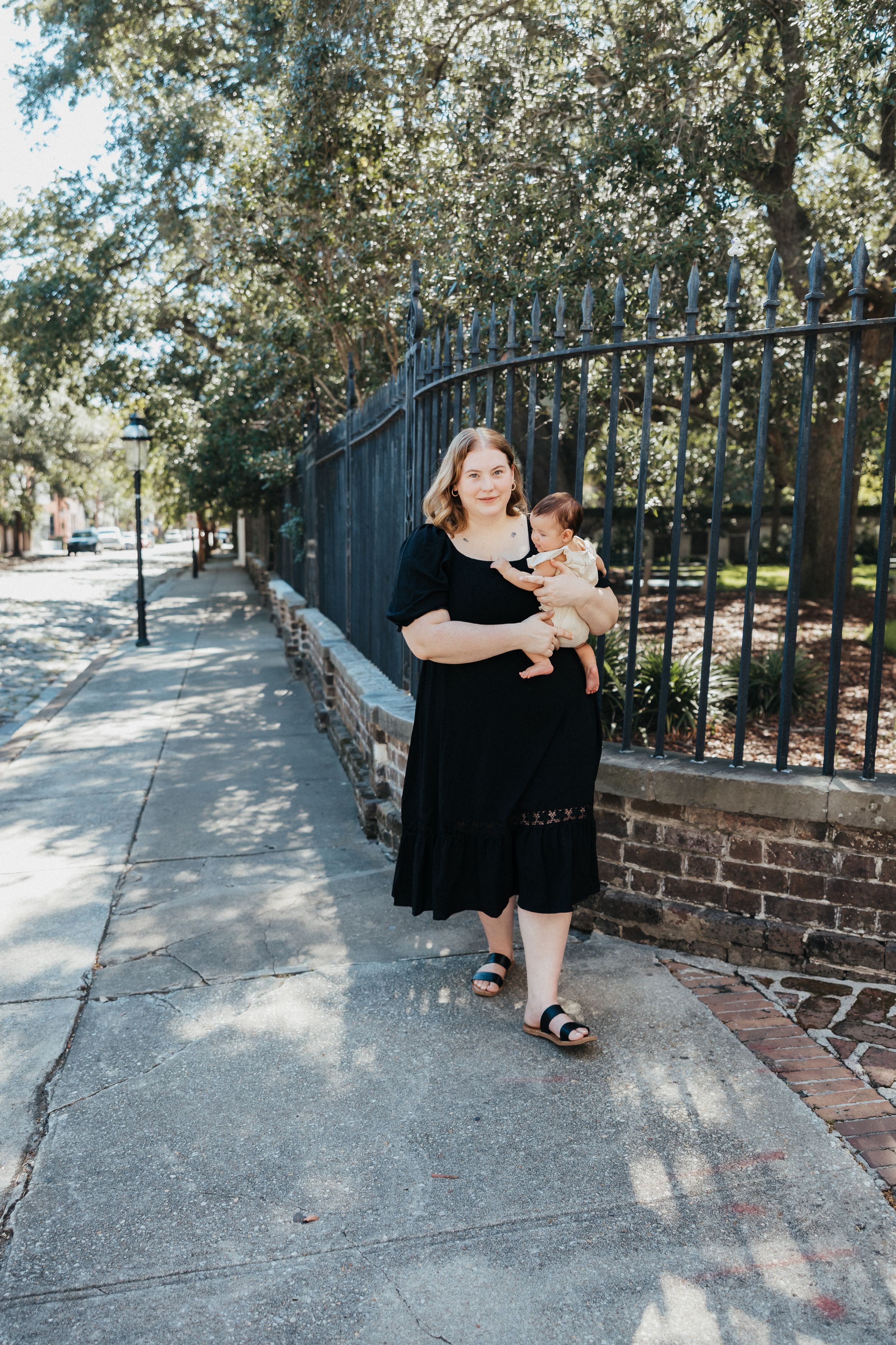 A woman in a black dress stands with a baby next to a wrought iron fence on a sunny sidewalk.