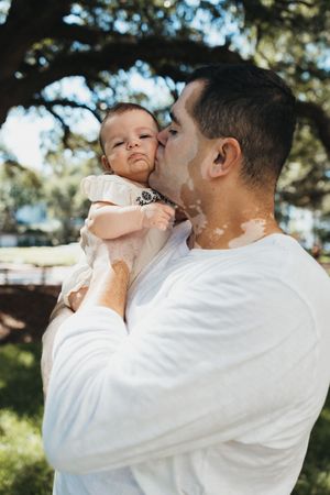 Sweet interaction between parent and baby in white clothing outdoors on a sunny day.