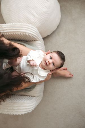 A child in white clothing lies on a cream-colored cushion while looking up at the camera in a minimalist setting.