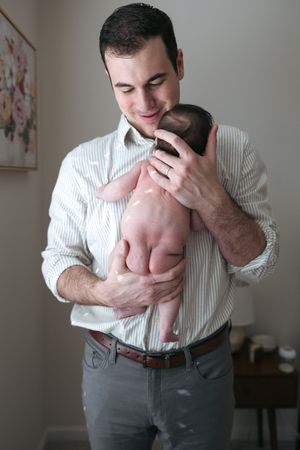 Man in striped shirt holding and comforting a small baby while standing in a room.