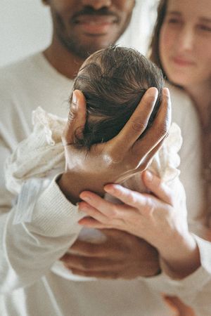Gentle hands cradle a newborn baby wrapped in white knitwear as parents share an intimate moment.