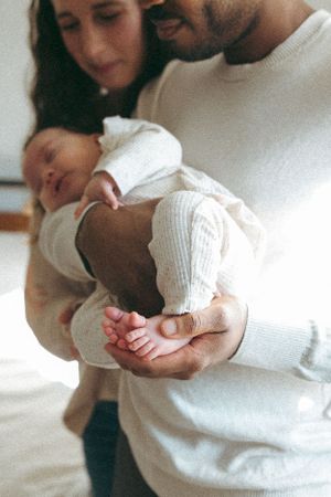 Parents in white sweaters tenderly hold their sleeping newborn baby, gently cradling tiny feet in an intimate moment.