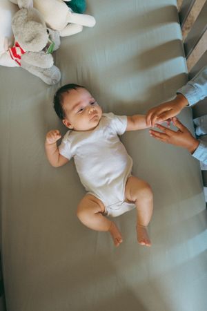 Baby wearing white onesie lying in crib with plush toys while someone gently holds their hand.