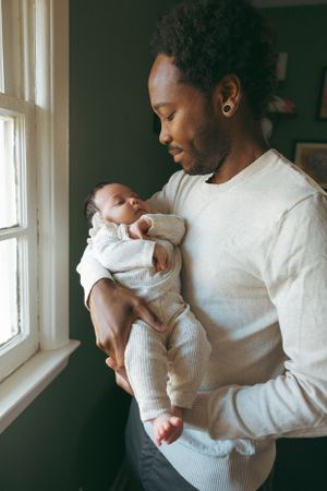 Adult in white sweater tenderly holds infant by window with natural light streaming in.