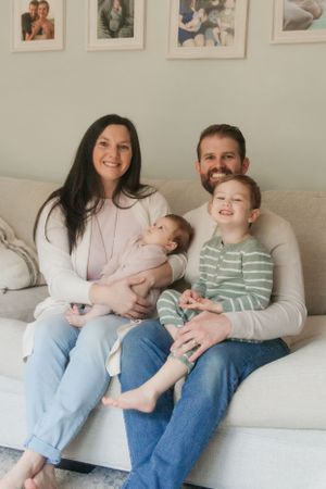 Happy family of four sitting together on a cozy grey couch with family photos displayed on wall.