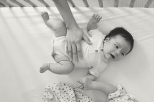 Baby lying in white crib wearing white onesie while adult hands reach down to change diaper.