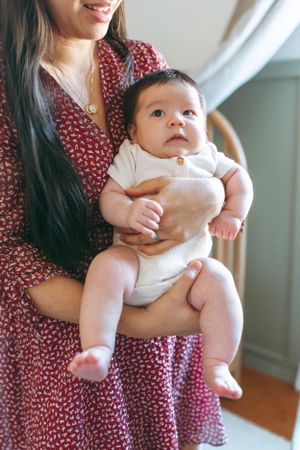 Baby in white onesie being held by person wearing floral red dress.