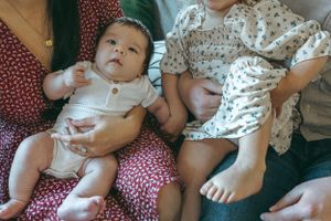 A baby wearing a white romper sits on a floral print cushion beside another person in a patterned outfit.