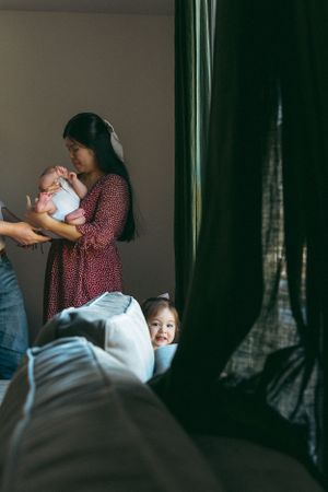 Moody indoor portrait of a mother holding a baby while another child peeks from behind furniture in dark lighting.