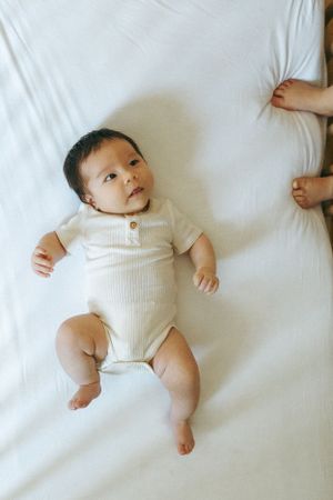 Baby wearing white ribbed onesie lies on white bedding with feet and arms outstretched.