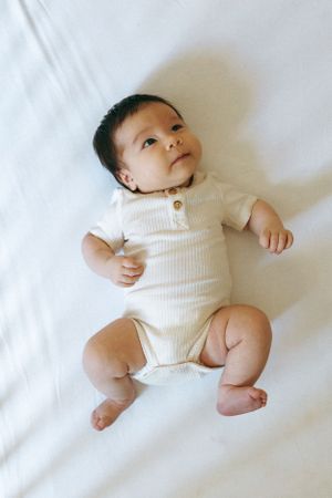 Baby wearing white ribbed onesie lies on white bedding and looks up with curiosity.