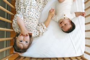 Two children lie on white bedding in a wooden crib, relaxing and playing together.